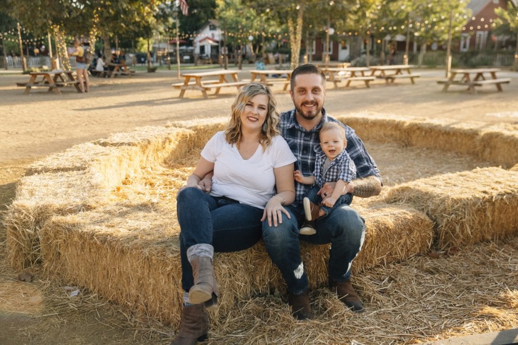 Julia, Nathan, and son, Archer Muzquiz, sitting on a hay bale in front of picnic tables, posing for a family photo.