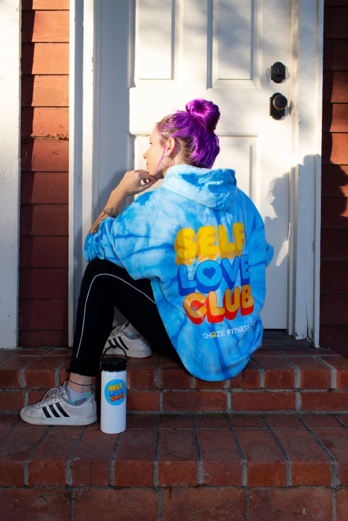 Woman in sitting in front of a doorway with our blue "Self Love Club" hoodie and a water bottle covered in our Self-Care Sticker Pack