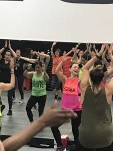 Instructor, Bettly Layman, in front of the Zumba Group Exercise class in an orange and pink Zumba shirt with her hands up in the air.