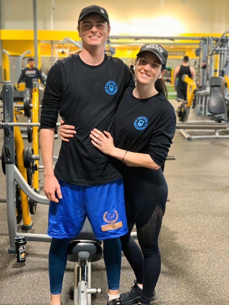 Conor on the left, Rissy on the right inside of Chuze Fitness near the weight machines. They are both wearing black hats, and black Chuze Fitness shirts with a blue logo from the Origin apparel collection.